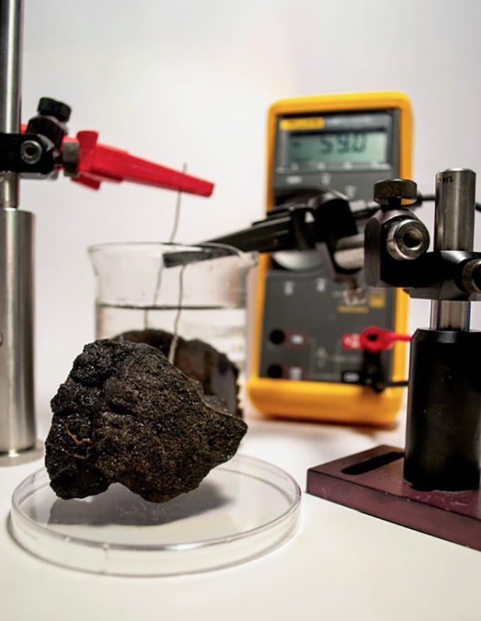 In a laboratory, wires held by clamps emerge from a metal-rich piece taken from the seabed in an experiment testing the electrical potential of the joint.